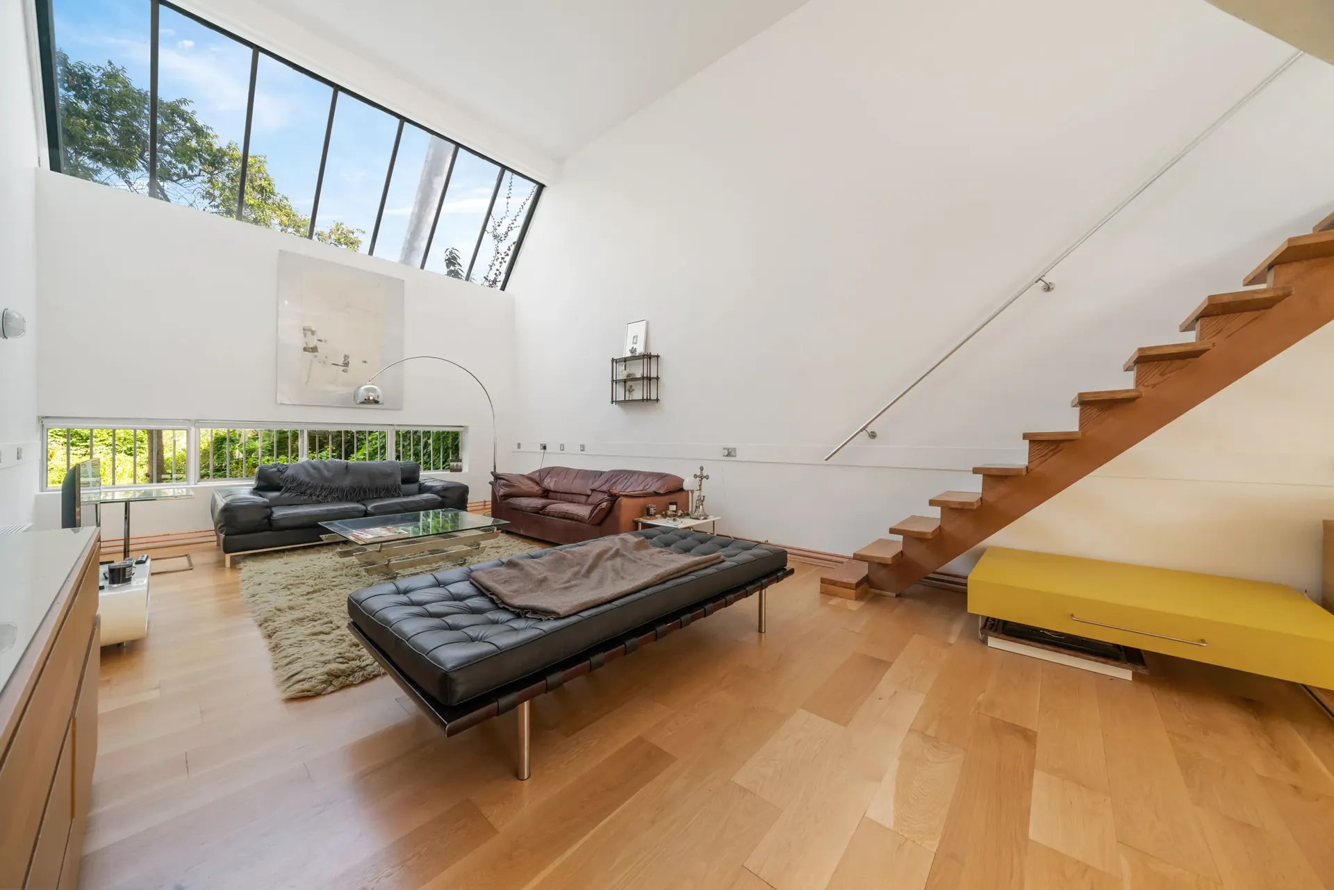 Living area with high-level glazing, tree views and a timber staircase to the bedroom